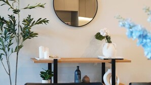 A room with a round mirror on the wall, a wooden table with a vase of flowers, and a potted plant.