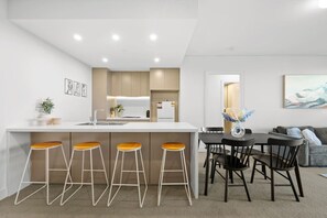 A modern kitchen with a central island, bar stools, a dining table with chairs, and a living area with a couch and artwork. - Luxury 2BR Oasis – Panoramic CBD & Park Views (Sydney Olympic Park)