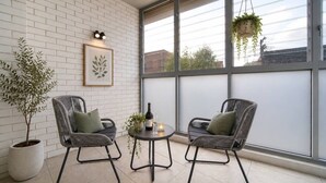 En balkon med to fletstole, et lille bord med en flaske og et glas, en potteplante og et indrammet kunstværk på væggen.