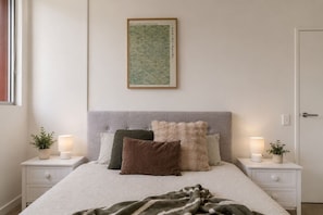 Une chambre à coucher comprenant un lit, deux tables de chevet, une petite table, une œuvre d’art encadrée et deux plantes en pot.