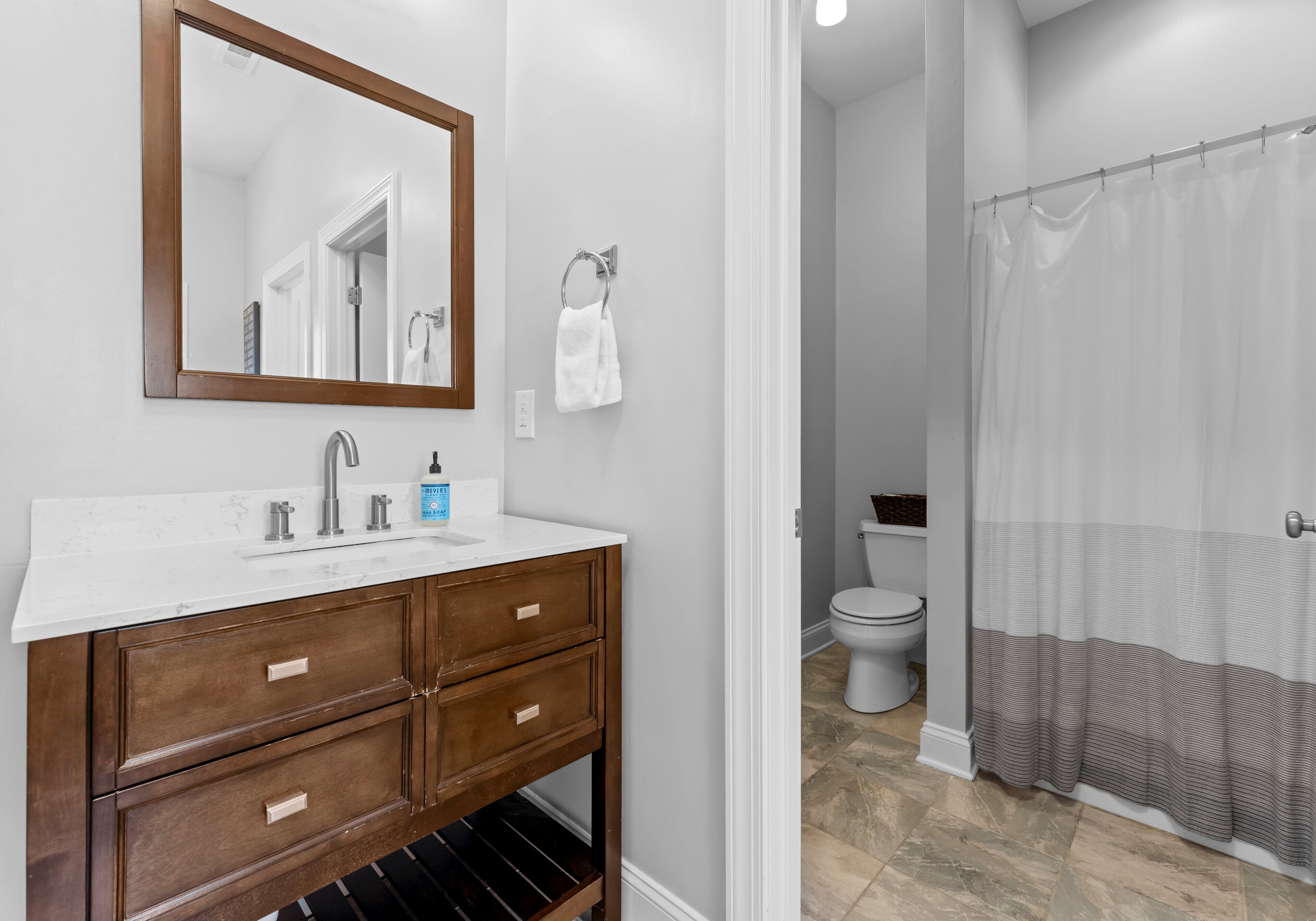 Un bagno con un lavandino bianco, una cassettiera in legno, uno specchio, un porta asciugamani, un water e una tenda per la doccia.