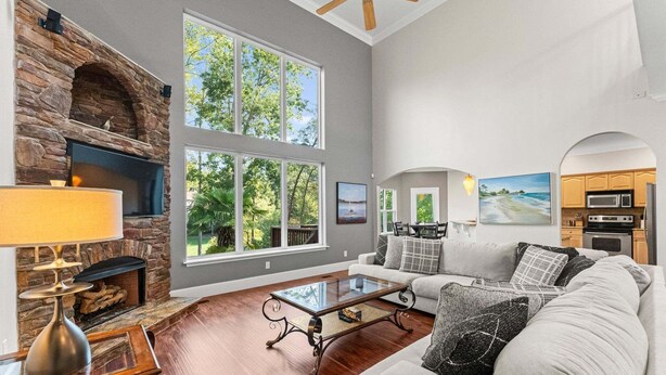 A living room with a stone fireplace, large windows, a ceiling fan, a sofa, and a kitchen area. - Lakeside Landing | Game Room | Sleeps 16 (Mooresville)