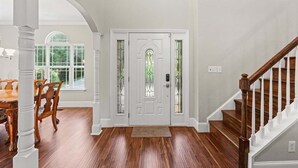 A foyer with a wooden staircase, a white door with stained glass, and a dining area with a table and chairs. - Lakeside Landing | Game Room | Sleeps 16 (Mooresville)