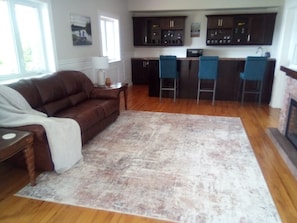 A living room with a brown sofa, a coffee table, a rug, and a kitchen area with dark cabinets and bar stools.