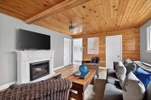 A living room with a fireplace, a television, a ceiling fan, and a view of the outdoors.