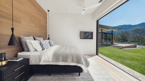 A modern bedroom with a large bed, a nightstand, a lamp, and a view of a lush garden and mountains. - Mountain Springs - Kangaroo valley (Barrengarry)