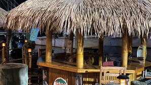 Uma área de estar ao ar livre com telhado de palha, pilares de bambu, uma placa com o nome 'Cavin Tiki' e vista para um cais ao fundo.