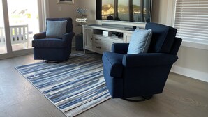 Uma sala de estar com dois poltronas azuis, uma televisão de tela plana grande, um armário de madeira e um tapete listrado.