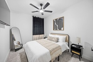 Una camera da letto con un letto, un comodino con una lampada, una ventola a soffitto, uno specchio e un'opera d'arte appesa al muro.