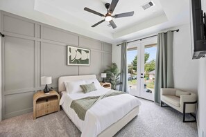 Ein Schlafzimmer mit Bett, Nachttisch, Deckenventilator, Fenster mit Vorhängen und Blick ins Freie.