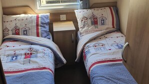 A compact RV interior with two beds, a window with blinds, and a small table. - 3-bed Caravan, Sleeps 8 With Enclosed Decking (Fleetwood)