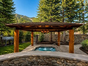 Una piscina de piedra con una pérgola de madera sobre ella, rodeada por un muro de piedra y una zona vallada con una estatua.