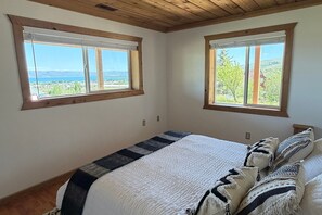 A bedroom with a large bed, two windows with wooden frames, and a view of a landscape.