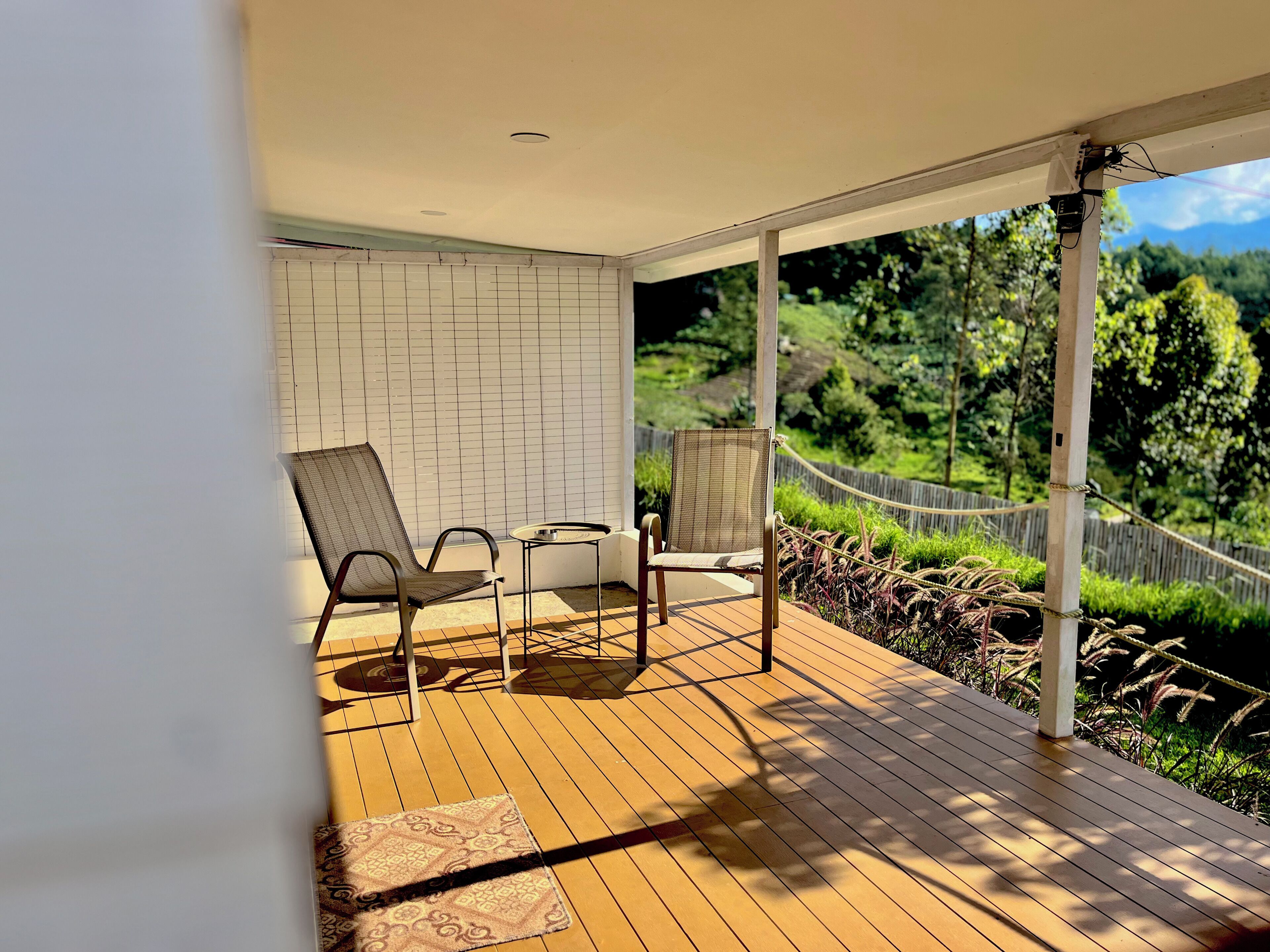 A wooden deck with two chairs and a small table, a white brick wall, and a view of greenery.