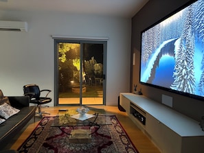 Moderni olohuone, jossa on suuri litteänäyttöinen televisio, lasinen sohvapöytä, sohva ja liukuovi, joka johtaa ulkotilaan.
