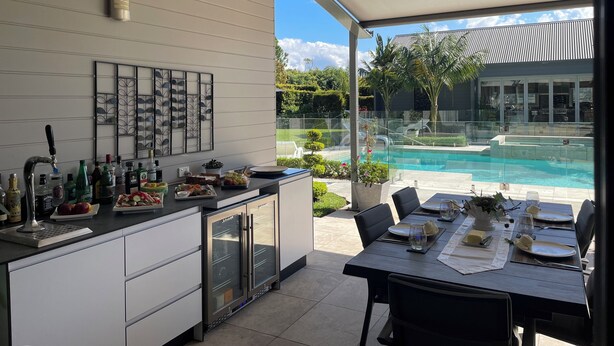 A modern kitchen with a built-in refrigerator, a bar area with bottles, and a dining table set for two with a view of a swimming pool. - Luxury Paradise Country Suite, Nature Retreat and Private Chef Fine dining (Duffys Forest)