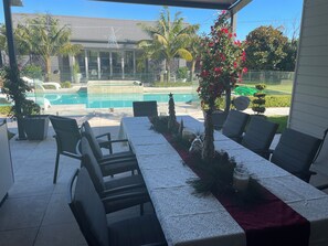 Un patio exterior con mesa lista para comer, sillas, una piscina y una casa al fondo.