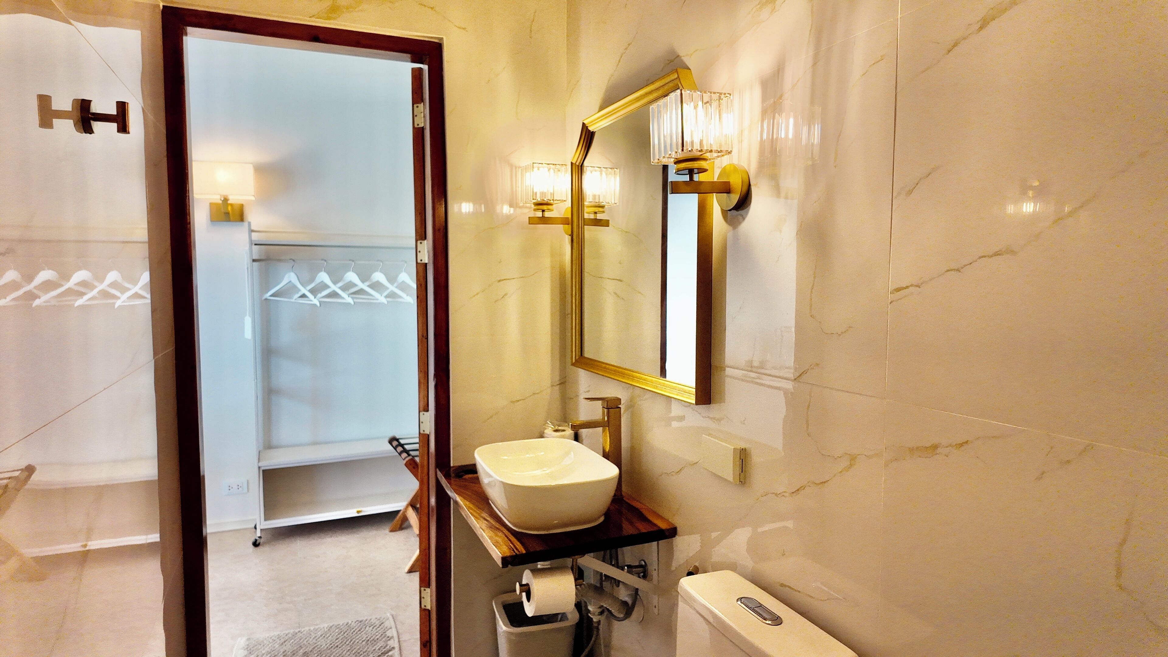 Un baño de hotel con pared de mármol, un lavamanos blanco, un espejo con marco dorado y un perchero para toallas.
