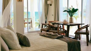 A bedroom with a bed, a table, chairs, a potted plant, and a view of the ocean.
