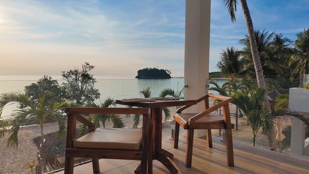 A wooden table and chairs on a balcony overlooking a beach with palm trees and a view of the sea.