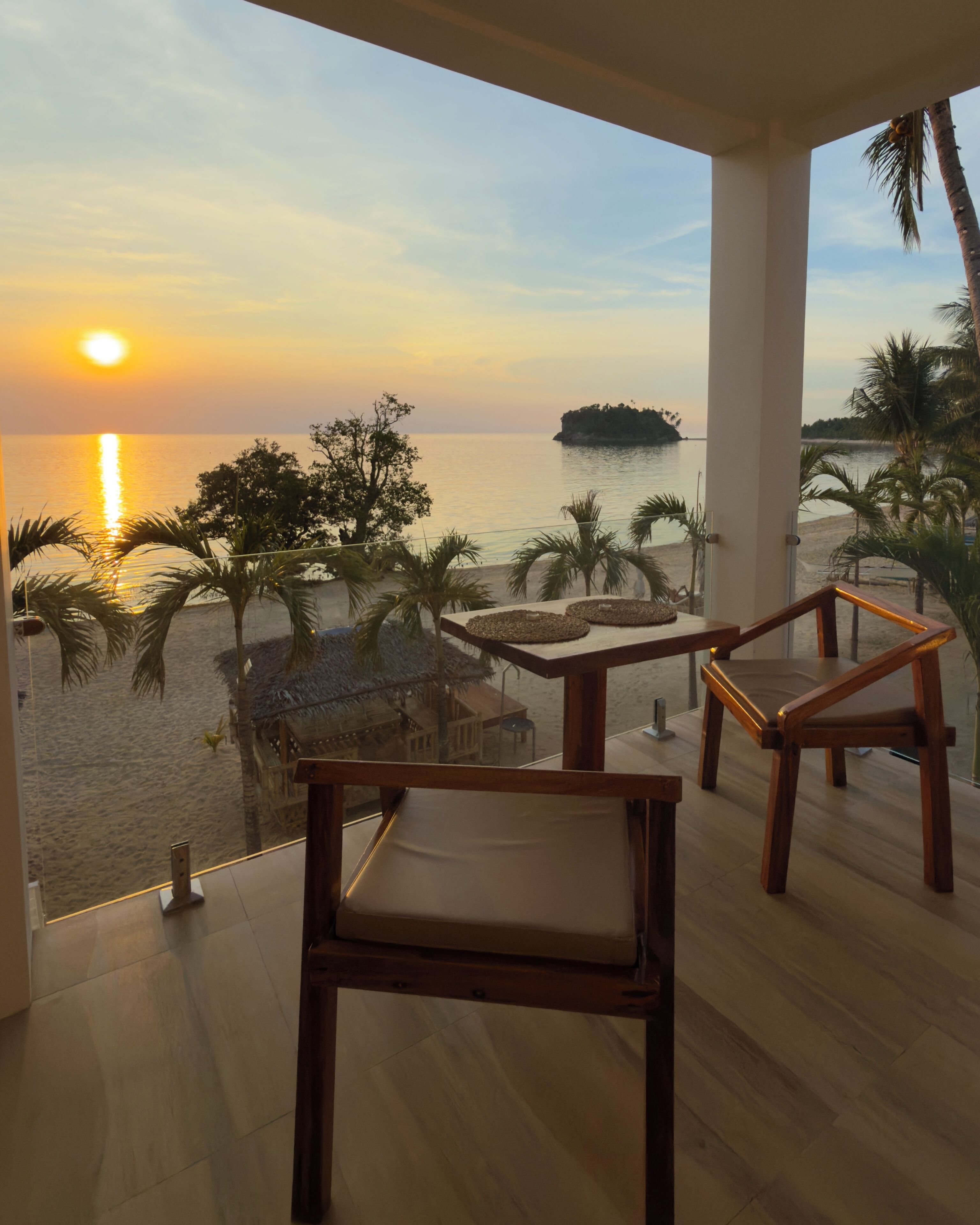 Un balcon avec deux chaises en bois, une balustrade en verre et une vue sur le coucher de soleil sur la mer, avec des palmiers et une petite île.