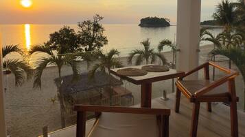 Un balcon avec deux chaises en bois, une balustrade en verre et une vue sur le coucher de soleil sur la mer, avec des palmiers et une petite île.