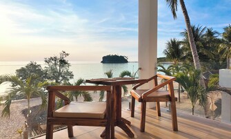 Una mesa y sillas de madera en un balcón con vistas a una playa, palmeras y al mar.
