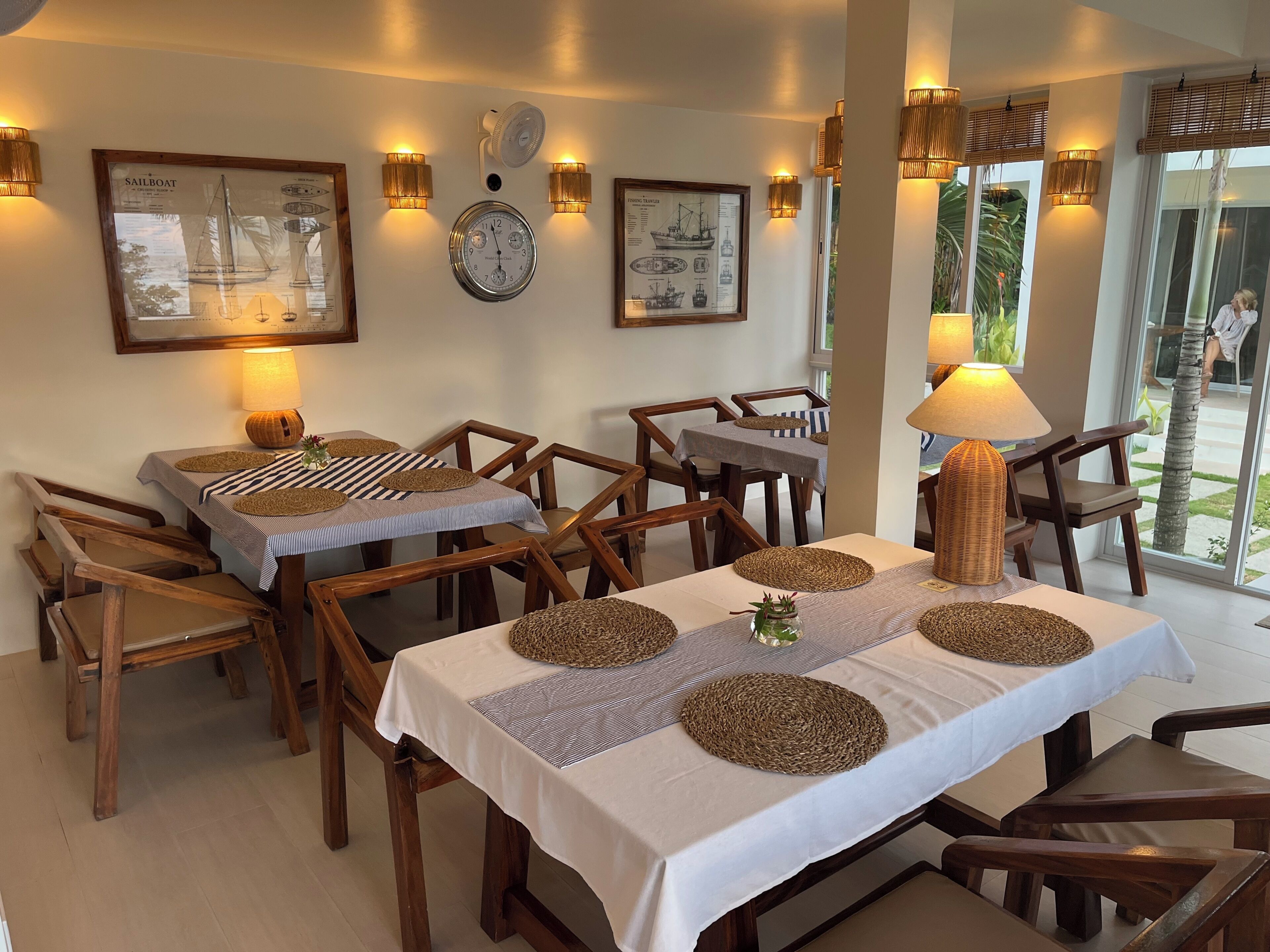 A dining area with wooden tables and chairs, framed nautical artwork on the walls, and a view of the outdoors through large windows.