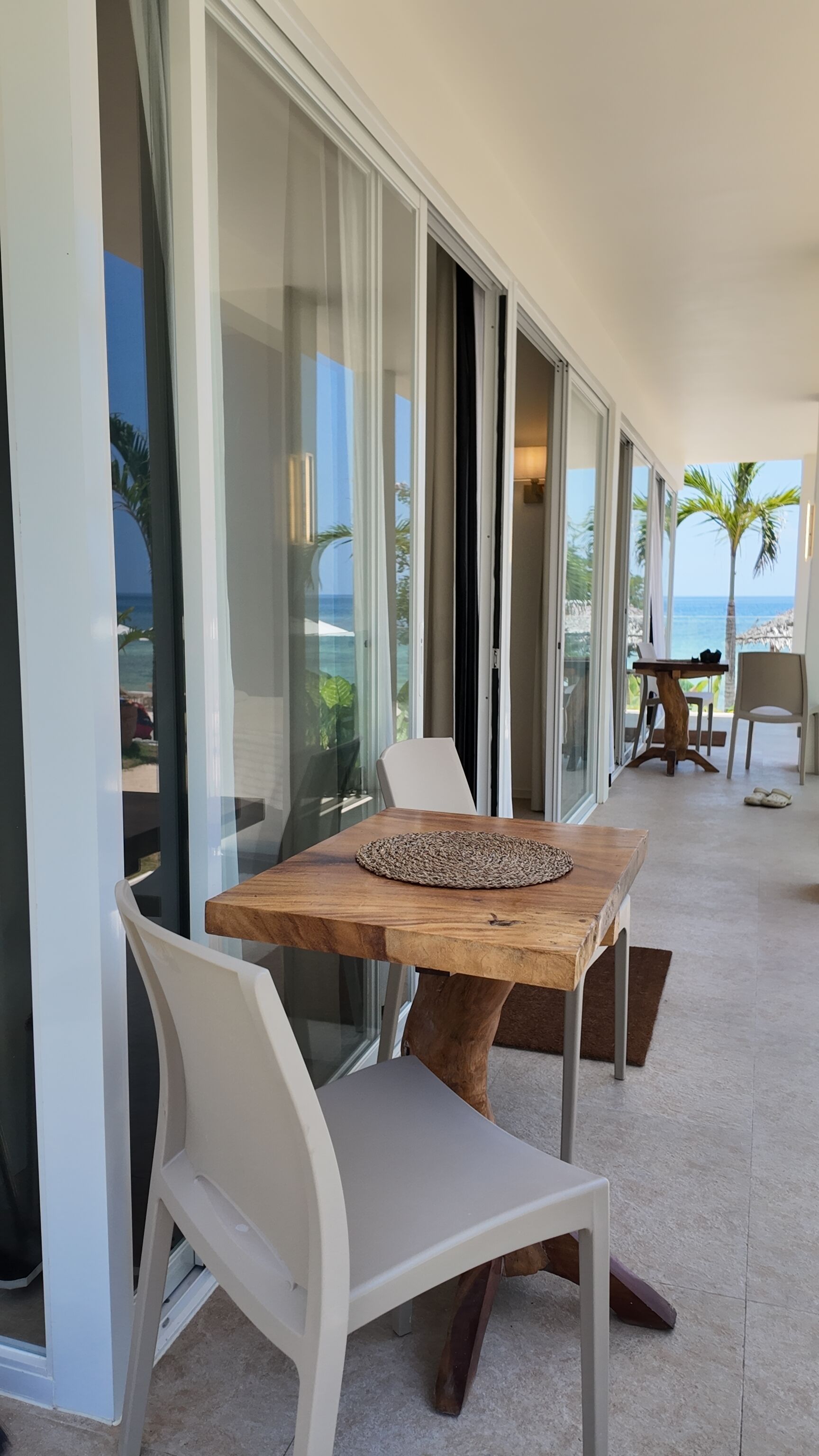 Ein zeitgenössischer Essbereich im Freien mit einem Holztisch und weißen Stühlen, mit Blick auf einen Strand und Palmen.