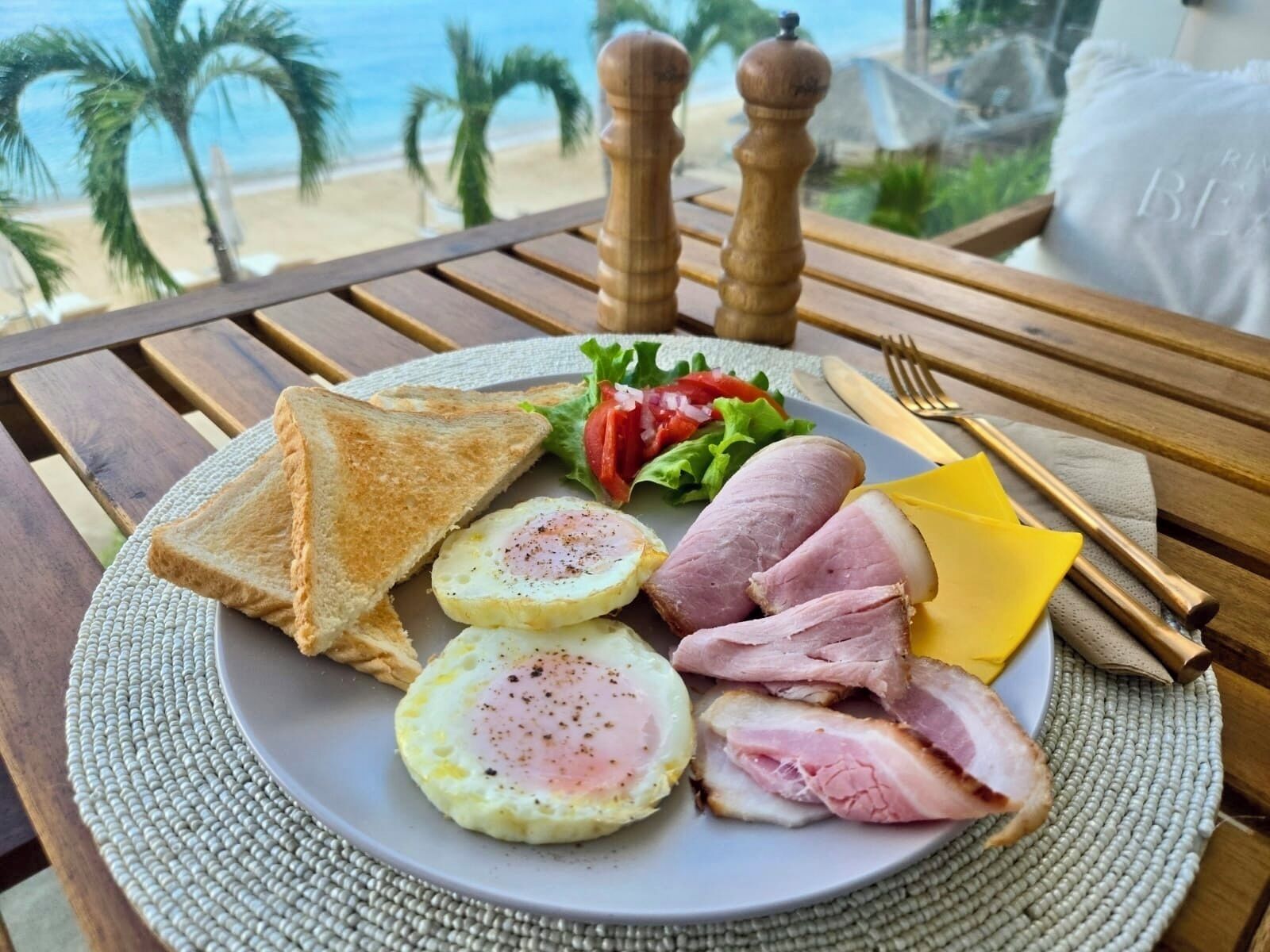 A breakfast plate with toast, eggs, ham, cheese, and salad on a table with a view of the beach.