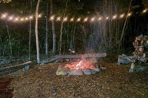 A campfire with a metal grill, surrounded by logs and rocks, with string lights overhead.