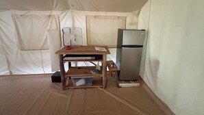 Una habitación con toldo, una mesa de madera, un ventilador, un refrigerador y un contenedor de plástico.