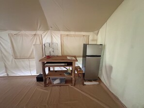 Une piÚce aménagée avec une tente, une table en bois, un ventilateur, un réfrigérateur et une boßte en plastique.