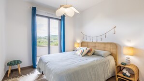 A bedroom with a bed, a nightstand, a hanging light fixture, a window with curtains, and a view of greenery outside. - Fiore di Macchja - À 5 min à pied de la plage (Albitreccia)