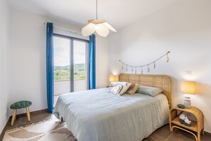 A bedroom with a bed, a nightstand, a hanging light fixture, a window with curtains, and a view of greenery outside. - Fiore di Macchja - À 5 min à pied de la plage (Albitreccia)