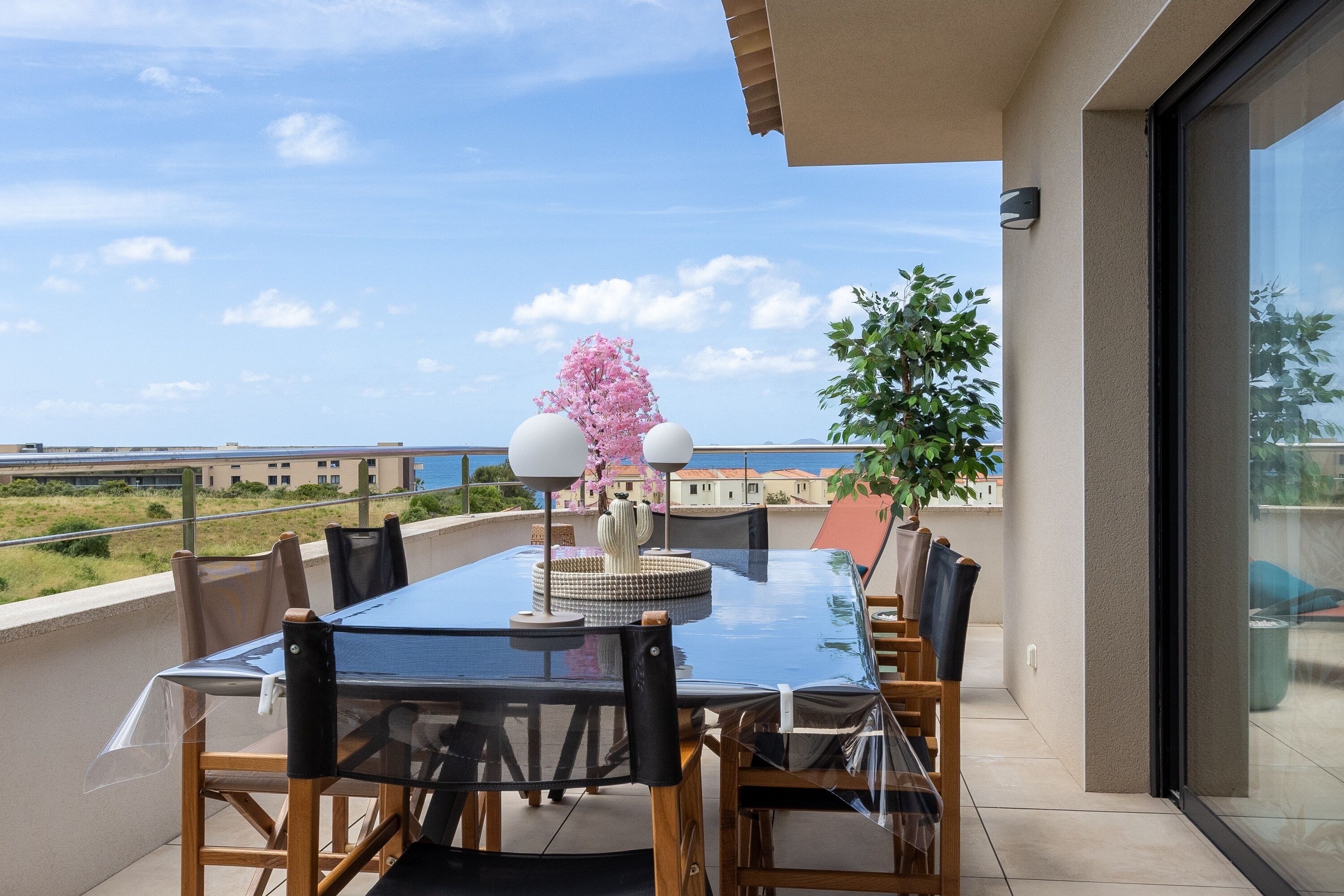 Un balcon avec une table et des chaises en verre, une plante en pot et une vue sur la mer.
