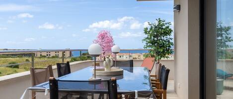 Un balcon avec une table et des chaises en verre, une plante en pot et une vue sur la mer.