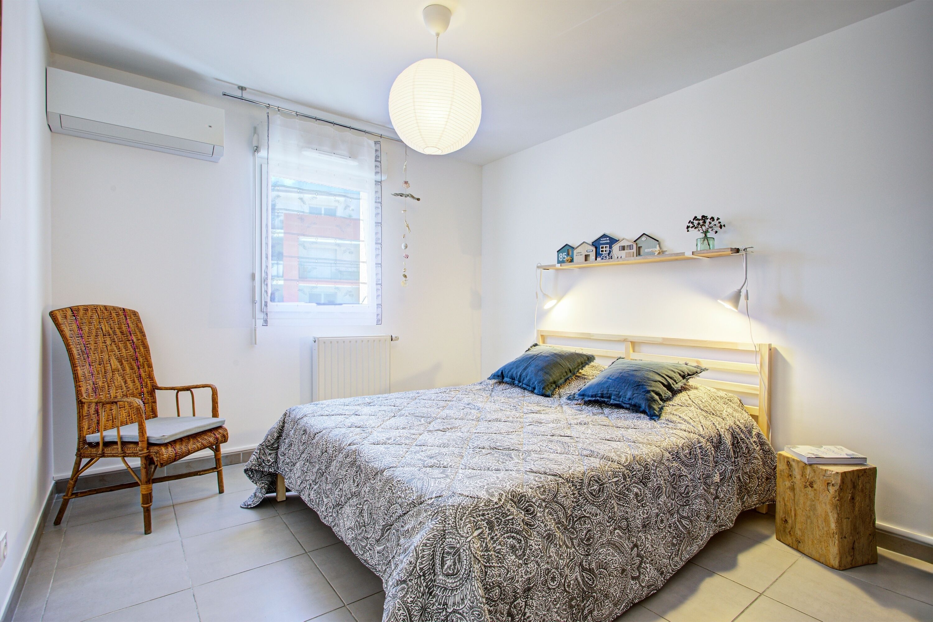 A bedroom with a bed, a chair, a bedside table, a shelf with decorative items, and a window with curtains.