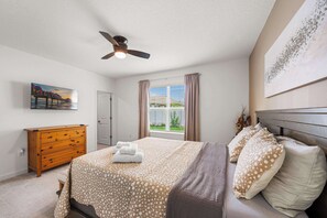 A bedroom with a bed, a ceiling fan, a window with curtains, a dresser, and artwork on the wall.
