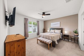 A bedroom with a bed, a bench, a desk, a ceiling fan, a TV mounted on the wall, and a window with curtains.