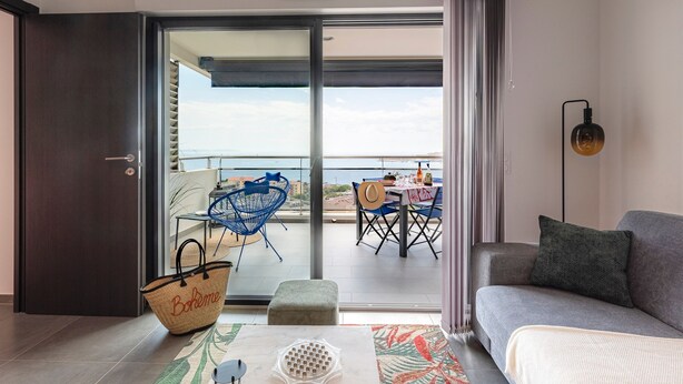 A modern living room with a grey sofa, a patterned rug, a large window with a view of the sea, and a dining area with blue chairs and a table. - Luminella - Avec vue sur la mer (Ajaccio)