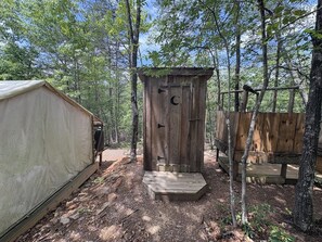 Un petit bâtiment en bois rustique, servant de toilettes, avec une découpe en forme de croissant de lune, entouré d’arbres.