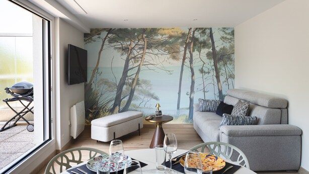 A living room with a sofa, a dining table with plates and glasses, a baby stroller, and a mural of trees. - Da Viken - Accès direct à la plage (Dinard)
