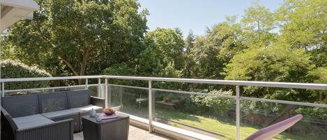 A balcony with a glass railing, outdoor furniture, and a view of trees and greenery.