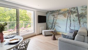 A modern living room with a large window, a flat-screen TV, a grey sofa, and a dining area with a table set for two. - Da Viken - Accès direct à la plage (Dinard)