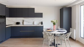 A modern kitchen with dark cabinetry, a marble countertop, and a dining area with white chairs and a table set for two. - Da Viken - Accès direct à la plage (Dinard)