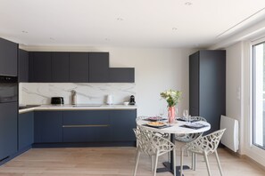 Een moderne keuken met donkere kasten, een marmeren aanrecht en een eethoek met witte stoelen en een tafel voor twee personen.