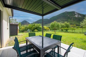 En overdækket terrasse med et bord og stole, udsigt til et bjerg og en indhegnet have.