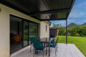 En moderne terrasse med et glasbord og stole, en skydedør der fører til et indre rum, og et bjerg i baggrunden.