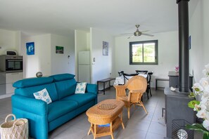 En stue med sofa, fletstole, et bord, en loftsvifte, et vindue og en brændeovn.